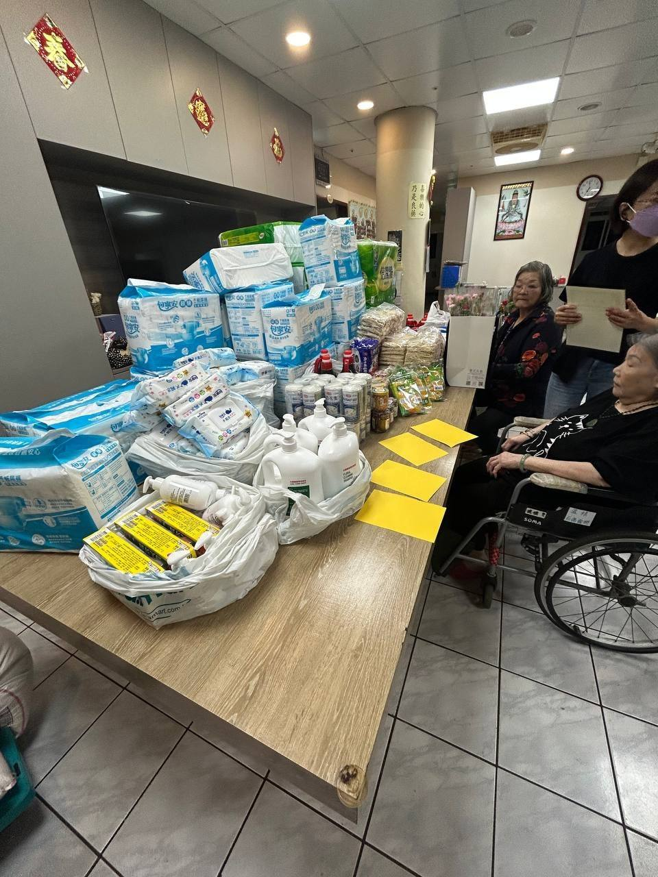 桌面擺放紙品、清潔用品與食品，旁邊有人們相聚